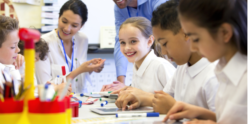 A group of school children can be seen working on digital tablets, two teachers can be seen behind them helping and supervising