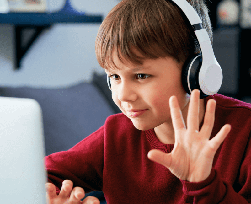 A child with headphones waving at a laptop screen.