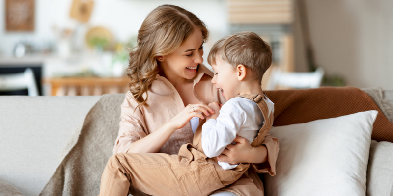 Delighted adult woman smiling and tickling laughing boy while chilling on sofa on weekend day at home together