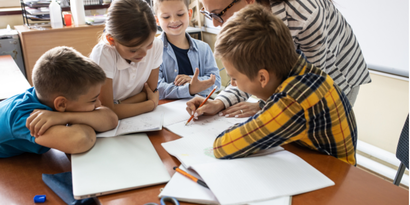 Female teacher helps school kids to finish they lesson.They sitting all together at one desk.