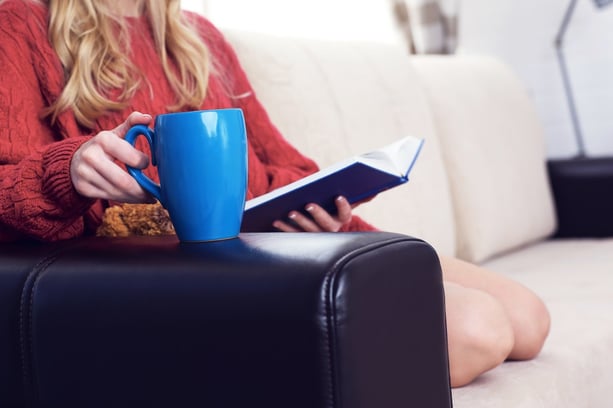 Middle section of young woman at home sitting on the couch relaxing in her living room reading book and drinking coffee or tea from blue cup.