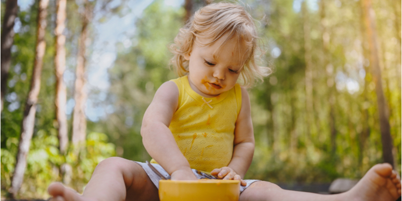 Little funny cute blonde girl child toddler with dirty clothes and face eating baby food