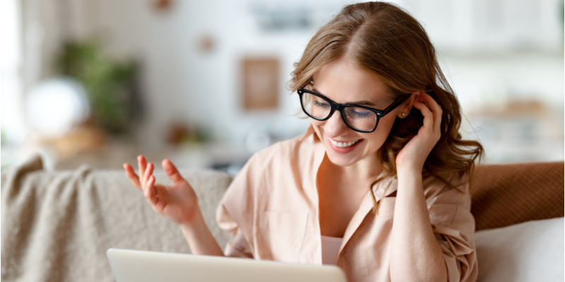 Positive young female in casual clothes and glasses sitting on couch at home and talking on video chat