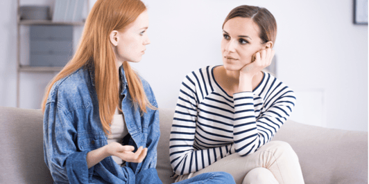 Teenage girl and a woman sitting on a gray couch and talking about problems in a bright, modern living room interior