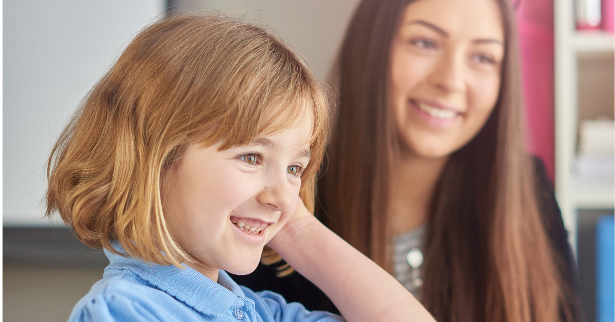 Image of a girl in school uniform using a computer, with a member of teaching staff sitting beside her. Both are smiling at the computer screen whilst the girl completes an activity.. 